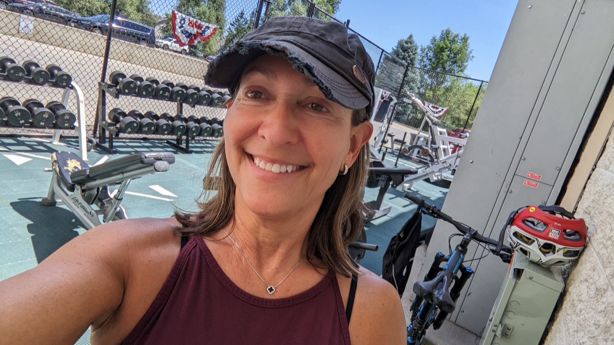 Alisa smiling at the outdoor weight floor, cap on, dumbbell racks behind her and her mountain bike leaning against the wall