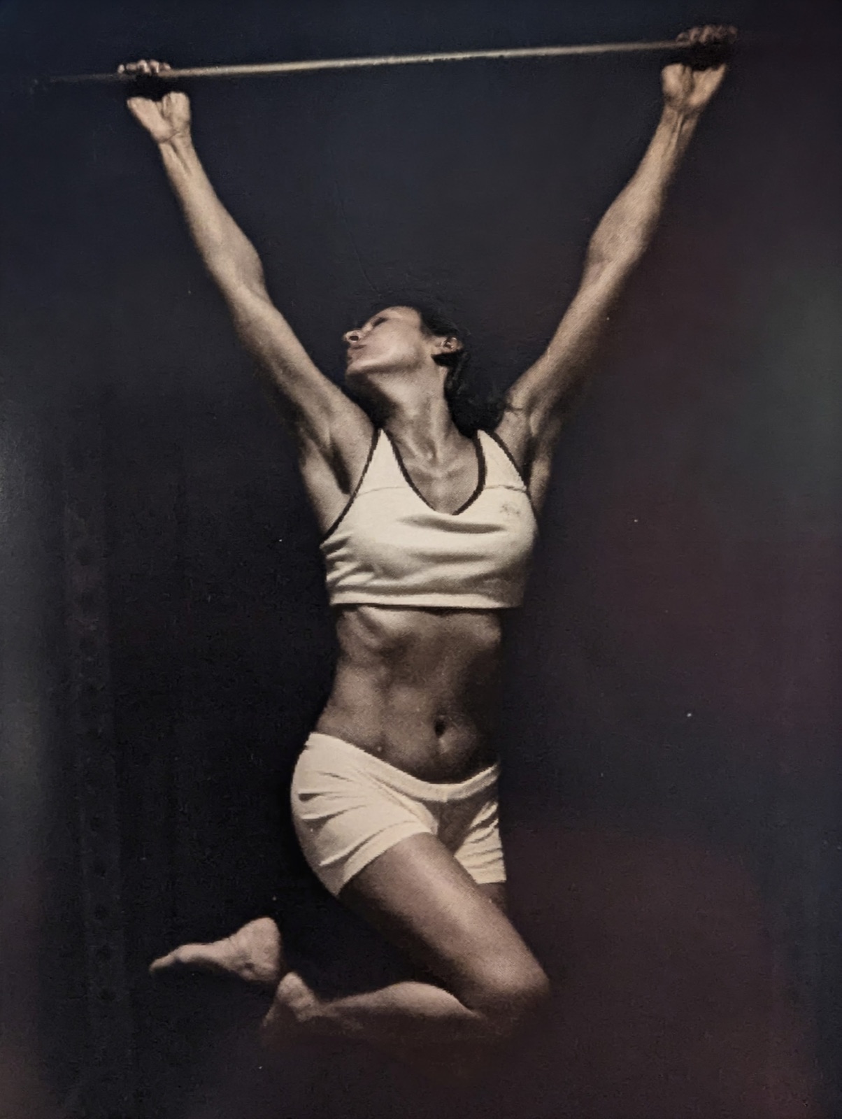 Studio photo of Alisa mid-rep on a pull-up bar, knees tucked, arms wide
